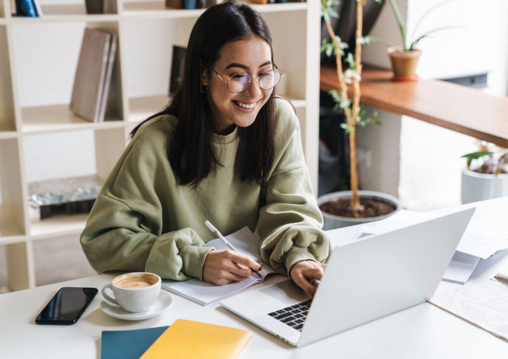 Eine Frau sitzt am Laptop und macht sich Notizen, sie schaut lächelnd auf den Bildschirm
